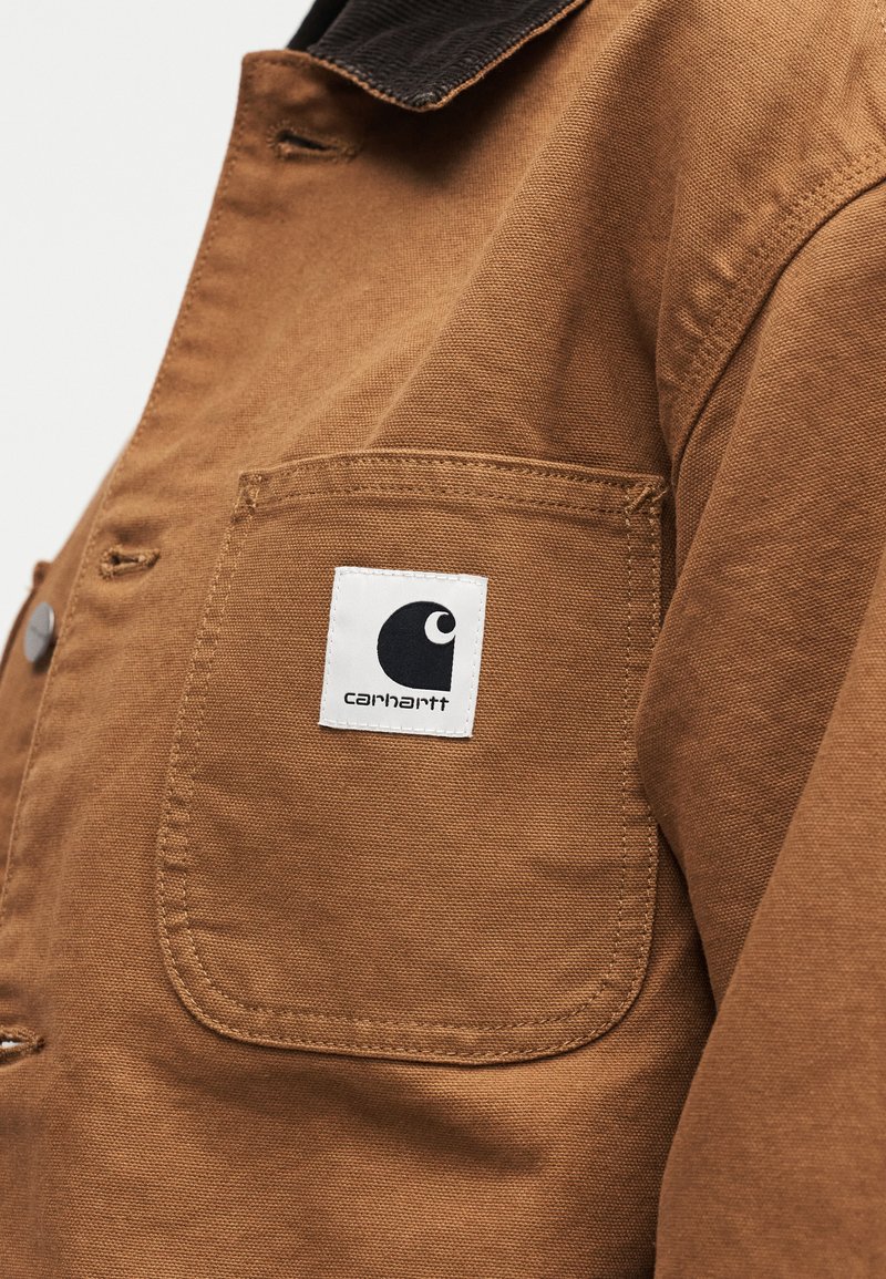 Tasca di una giacca in tela marrone con toppa logo Carhartt bianca che presenta una "C" stilizzata nera sull'area del petto.