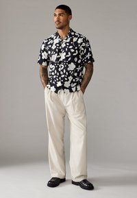 Korte mouwen zwarte shirt met een wit bloemenpatroon, gecombineerd met lichtbeige broeken en zwarte schoenen, met een ontspannen pasvorm en kraagontwerp.