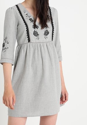 Grå klänning med 3/4 ärmar, V-ringning och blommig broderi. Har en samlad midja och texturigt tyg. Utsmyckad med svart spetskant.