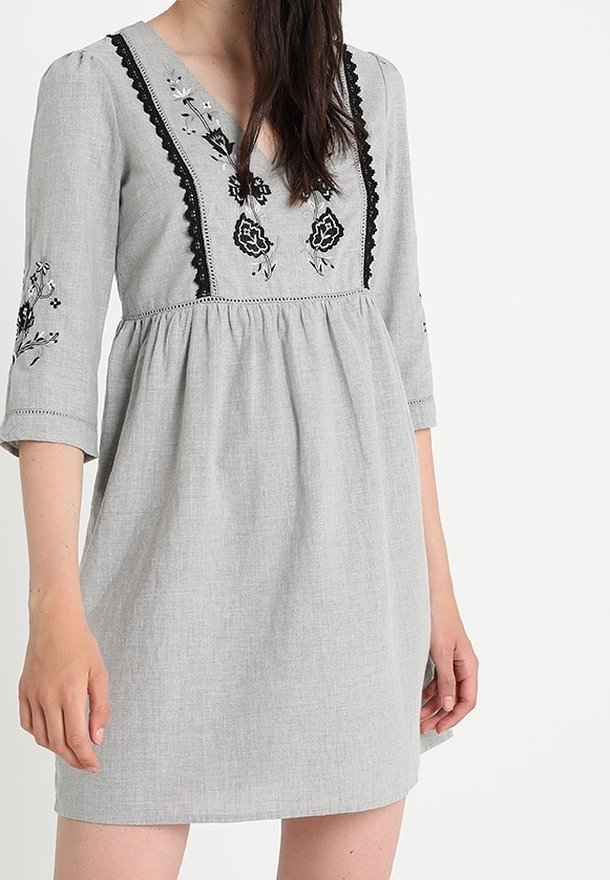 Grå klänning med 3/4 ärmar, V-ringning och blommig broderi. Har en samlad midja och texturigt tyg. Utsmyckad med svart spetskant.