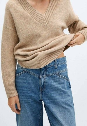 Person iført en beige V-hals sweater og blå højtaljede jeans, løfter let sweateren med den ene hånd.