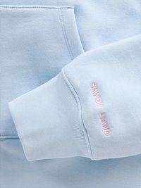 Sweatshirt azul claro com um bolso canguru e punhos de manga com ribanas. Apresenta texto bordado em rosa "SANDY LIANG" perto do punho.