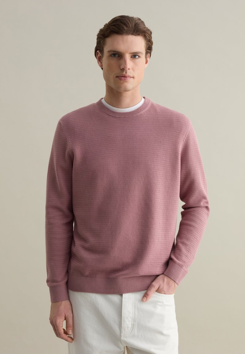 Pink knit sweater with a textured pattern, ribbed cuffs, and hem. Worn over a white T-shirt, displayed front-facing against a neutral background.