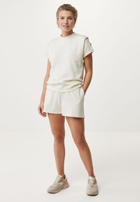 Weißes Kurzarmoberteil mit hochgerollten Ärmeln, kombiniert mit passenden Shorts. Hergestellt aus weichem Stoff und im lässigen Schnitt. Das Modell trägt beigetöne Sneaker.