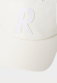 White baseball cap made of textured fabric featuring a prominent, stitched white letter "R" on the front. Adjustable strap at the back.