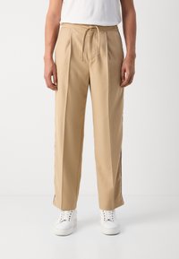 Beige skreddersydde bukser med elastisk liv og snor. Har sidestriper og løs passform. Kombinert med hvite joggesko.