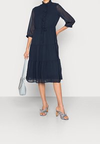 Námořnická modrá šifónová šaty s volánkovým límcem, detaily s knoflíky a vrstveným designem. Kombinováno s kostkovanými sandály na podpatku a světle modrou kabelkou.