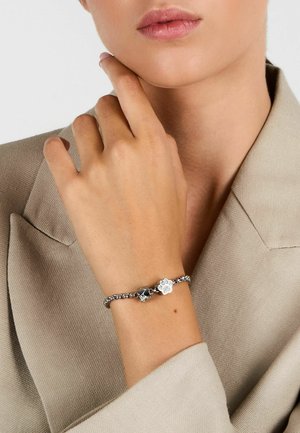 Braccialetto d'argento con design a catena, decorato con due charm: un'impronta di zampa e una silhouette di gatto. Indossato su un polso su un tessuto beige.
