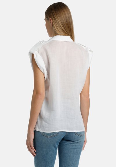 Camisa de lino blanca con mangas cortas dobladas, vista por detrás. Presenta un cuello clásico y un corte relajado, con una textura semitransparente. Se lleva con jeans azules.