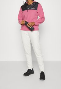 Pullover in pile rosa con pannello superiore nero e logo, abbinato a pantaloni della tuta grigi chiari e sneakers nere, mentre tiene in mano una bottiglia d'acqua.