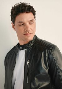 Czarna skórzana kurtka z kołnierzem stojącym, z przodem na zamek błyskawiczny i żebrowanym wykończeniem na ramionach. Nosi się ją na białej koszulce t-shirt na neutralnym tle.