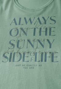 Top sin mangas de algodón verde menta con texto superpuesto en azul oscuro y acentos de strass. El texto incluye frases sobre positividad y autenticidad.