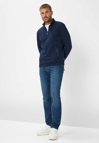 Navy blue zip-up pullover with stand collar, featuring side pockets, paired with blue jeans and white sneakers. Smooth texture, casual design.