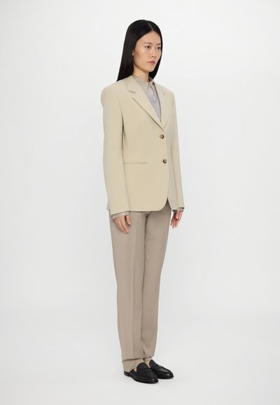Beige tailored blazer with two buttons, gray shirt underneath, beige tailored pants, and black loafers, standing on a white background.