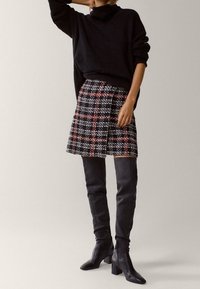 Zwarte oversized trui, rok met houndstooth-patroon in zwart, wit en rood, gecombineerd met zwarte leren kniehoge laarzen met hak.