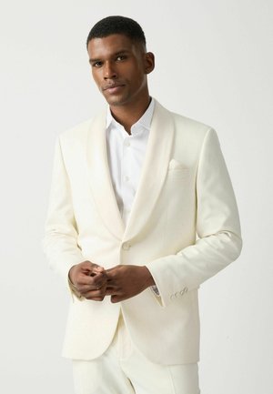 Man wearing a white tuxedo jacket with shawl lapel and white dress shirt against a plain white background.