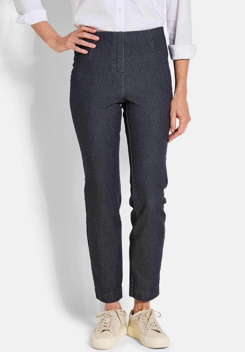Vrouw in donkere, aansluitende broek, wit overhemd met opgerolde mouwen en lichtbeige sneakers, staat met één hand op de heup.