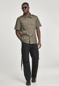 Camisa de manga corta color verde oliva con dos bolsillos frontales, combinada con pantalones cargo negros que tienen bolsillos laterales y zapatos negros.