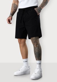 Black shorts with elastic waistband and adjustable drawstring. Smooth fabric, side pockets. Worn with white sneakers and patterned tattoos.
