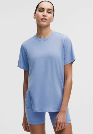 Mujer joven con camiseta de manga corta y pantalones cortos ciclistas a juego en azul claro, de pie contra un fondo liso y claro.