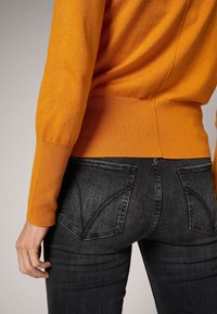 Orange knit sweater with a ribbed hem and long sleeves, paired with fitted black jeans featuring back pockets and subtle whiskering.
