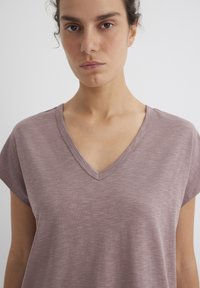V-Ausschnitt T-Shirt aus weichem, strukturiertem Mauve-Stoff mit kurzen Ärmeln und lockerer Passform. Detaillierte Naht entlang des Ausschnitts.