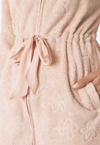 Lys pink morgenkåbe i blød, struktureret stof med blomsterprint, der har bindebånd i taljen og sidelommer for ekstra funktionalitet.