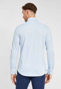 Camicia azzurro chiaro a maniche lunghe con colletto abbottonato, orlo arrotondato e polsini aderenti. Tessuto morbido con texture minima, mostrata da dietro.