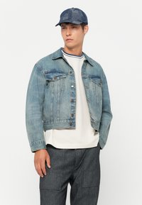 Joven vestido con una chaqueta de mezclilla azul, una camiseta blanca de cuello redondo con ribete azul, pantalones grises y una gorra de parches azul, de pie contra un fondo blanco.