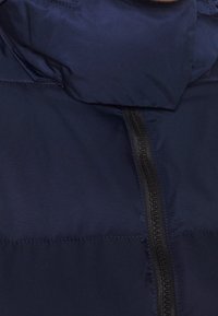 Marineblå pufferjakke med høj krave, lynlås og sømmet paneldesign. Glat tekstur med mat finish.