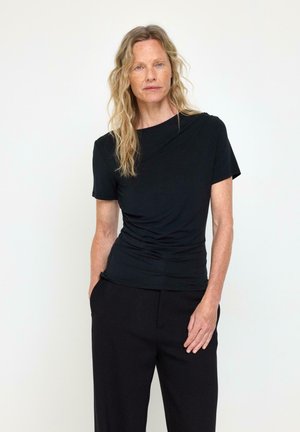 Donna con capelli chiari che indossa una maglietta nera aderente a maniche corte e pantaloni neri, in piedi davanti a uno sfondo chiaro e uniforme con le mani nelle tasche.