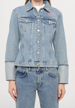 Femme portant une veste en denim bleu clair avec des boutons argentés et un jean taille haute assorti, debout devant un fond blanc uni.