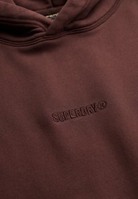 Felpa marrone con cappuccio realizzata in tessuto testurizzato, dotata di cappuccio e logo ricamato "SUPERDRY" sul fronte. Linee pulite e design minimale.