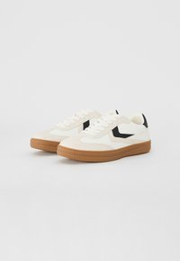 Sneakers z białą kanwasową cholewką, czarnymi akcentami i teksturowaną brązową gumową podeszwą. Posiadają klasyczny fason na sznurowania oraz okrągły nos.