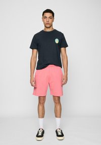 Marineblå t-shirt med korte ærmer og et farverigt logo på brystet, parret med lyserøde shorts med elastisk talje. Sorte sneakers med hvide såler.