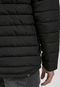 Sort pufferjakke med quiltet design, der viser vandret syninger, med en glat tekstur og et lille mærke nær kanten.