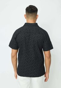 Chemise noire à manches courtes avec un motif géométrique texturé. Possède un col et un ourlet droit. Portée avec un pantalon couleur crème.