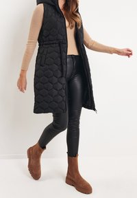 Schwarze gesteppte lange Weste mit Kapuze, olivgrünes geripptes Oberteil, glänzende schwarze Leggings und braune Wildleder-Stiefeletten mit Gummisohlen.
