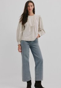 Lichtbeige gestreepte blouse met een geplooide halslijn, lange volumineuze mouwen; gecombineerd met lichtblauwe wijde jeans en zwarte enkellaarsjes.