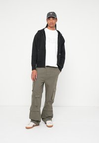 Μαύρο φούτερ με φερμουάρ πάνω από λευκό γραφικό t-shirt, ανοιχτό ελαιόχρωμο cargo παντελόνι με τσέπες και λευκά αθλητικά παπούτσια με καφέ σόλες, σε λευκό φόντο.