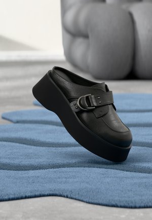 Μαύρο δερμάτινο mule πλατφόρμας με μεγάλο λουράκι, που βρίσκεται πάνω σε ένα υφασμάτινο, κυματιστό μπλε χαλί και θολό γκρι φόντο.