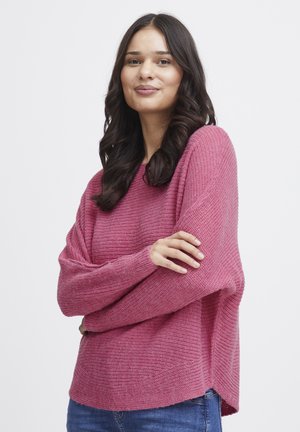 Pink knit sweater with a loose fit, textured design, wide sleeves, and a longer back hem. Worn with blue jeans.