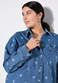 Denimhemd mit einem hellblauen Herzmuster, Knopfleiste vorne, zwei Brusttaschen und langen Ärmeln mit geknöpften Manschetten. Silbrige Ringdetails sichtbar.