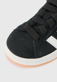 Black suede sneaker with a textured finish, featuring black laces and a white side stripe. Soft rubber sole with an orange accent.