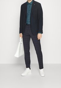 Marineblazer over een teal-gepatroneerde blouse, gecombineerd met marineblauwe geblokte broek en witte sneakers. Een witte tote bag maakt de outfit compleet.