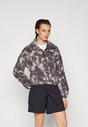 Felpa grigia a collo alto in camouflage, con zip a metà e logo sul petto, abbinata a pantaloni cargo neri.