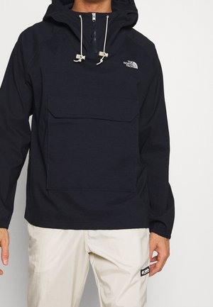 Mężczyzna ubrany w czarną kurtkę z kapturem The North Face z przednią kieszenią i białymi sznurkami, w zestawieniu z jasnymi spodniami.