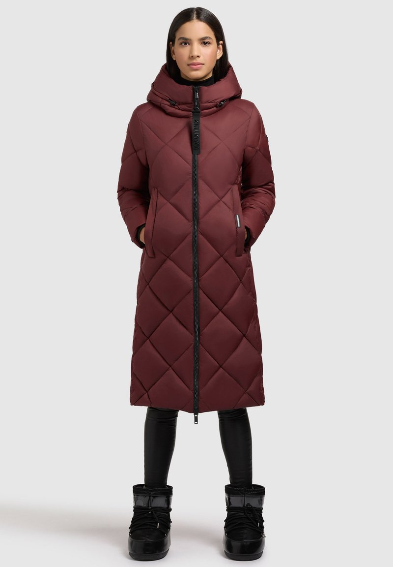 Bordeauxfarbener langer Puffermantel mit hohem Kragen, Rautenmuster, Frontreißverschluss und zwei Seitentaschen. Getragen über schwarzen Leggings und Stiefeln.