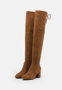 Botas de ante marrón sobre la rodilla con tacón cuadrado y un detalle de cordones en la parte superior. Textura suave con costuras mínimas visibles.