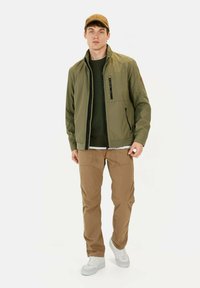 Olivgrüner Windbreaker mit Reißverschluss und Taschen, über einem dunkelgrünen Pullover getragen. Beigefarbene Hose und weiße Sneakers vervollständigen den Look.